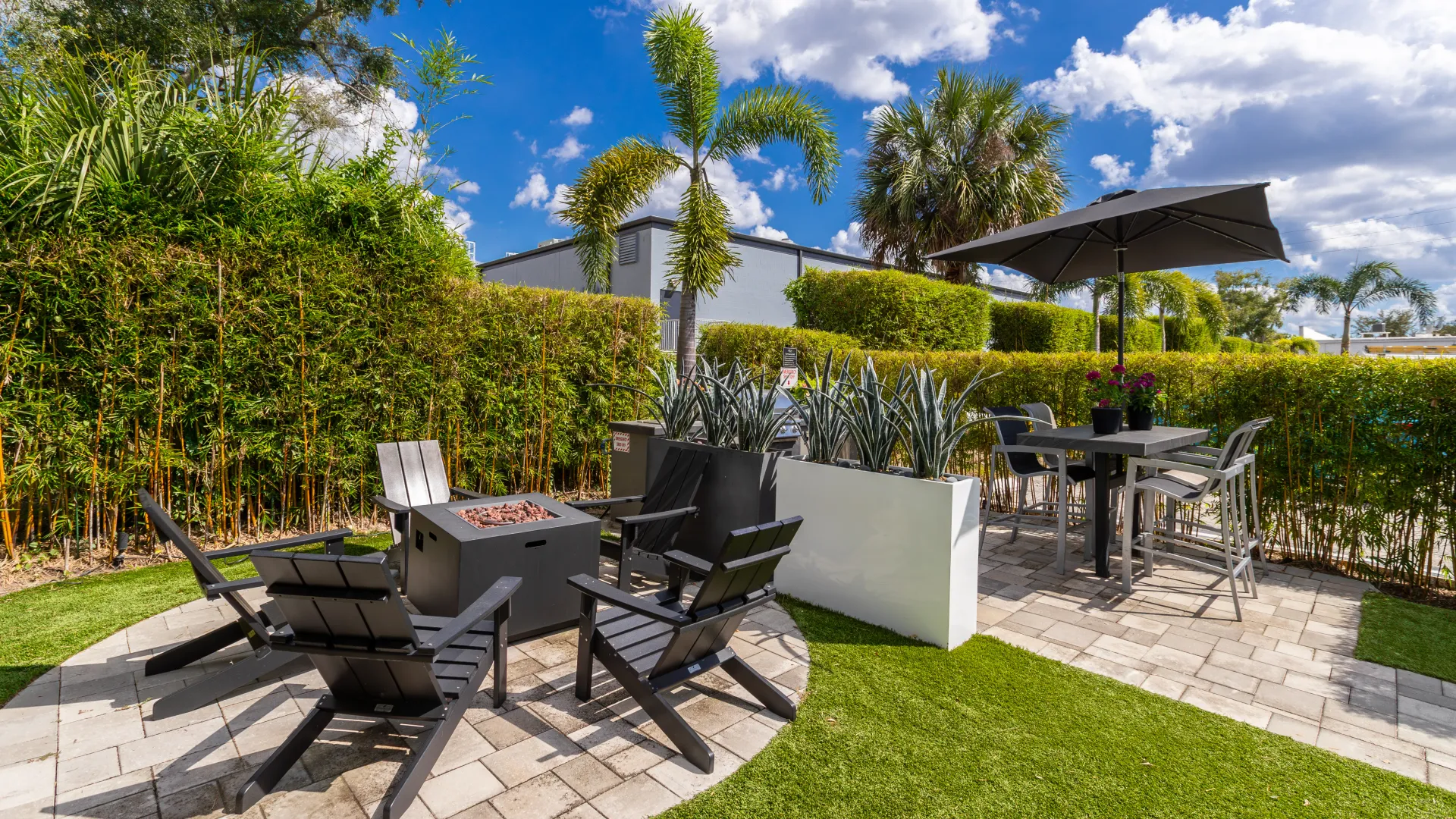 Outdoor lounge with a fire pit, dining table, and chairs, surrounded by lush greenery and palm trees.