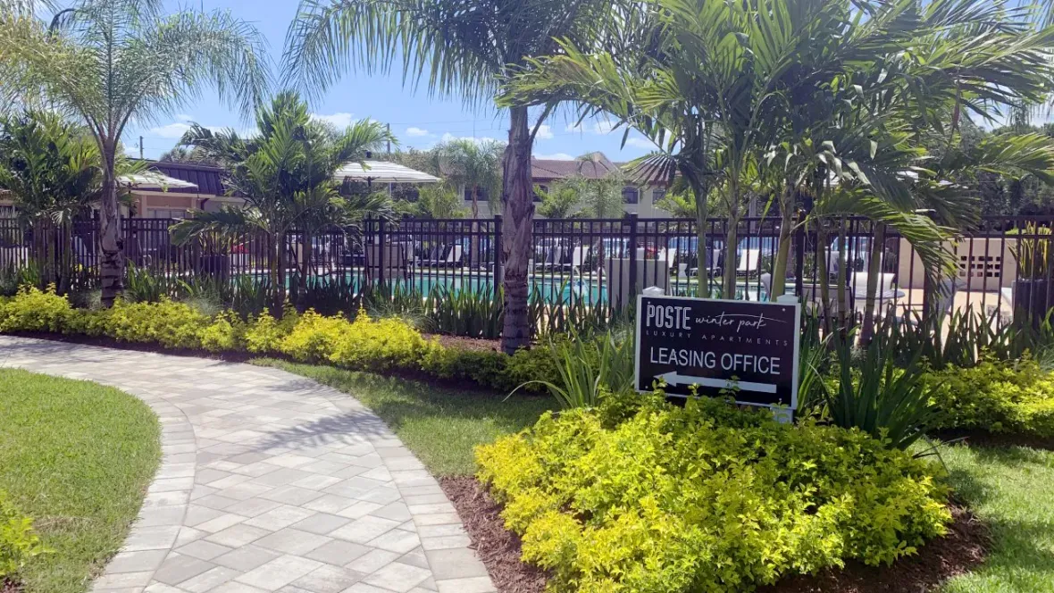 A paved pathway winding past a shimmering pool, flanked by tropical palm trees. A sign directs you to the leasing office, your gateway to a new home.