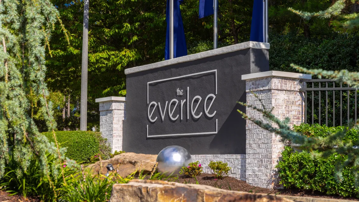 A majestic white-washed brick sign bearing The Everlee Apartments' logo against a backdrop of three vibrant blue flags, an inviting gateway promising warmth and comfort in every residence.