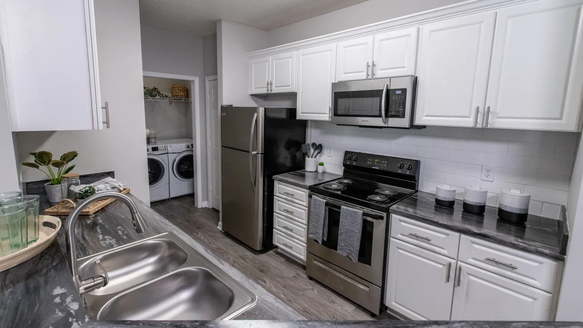A kitchen showcasing white shaker cabinets, a double sink, a stainless-steel appliance package, generously spacious black-fusion countertops, grey wood-like flooring, and a glimpse into the adjoining laundry room.