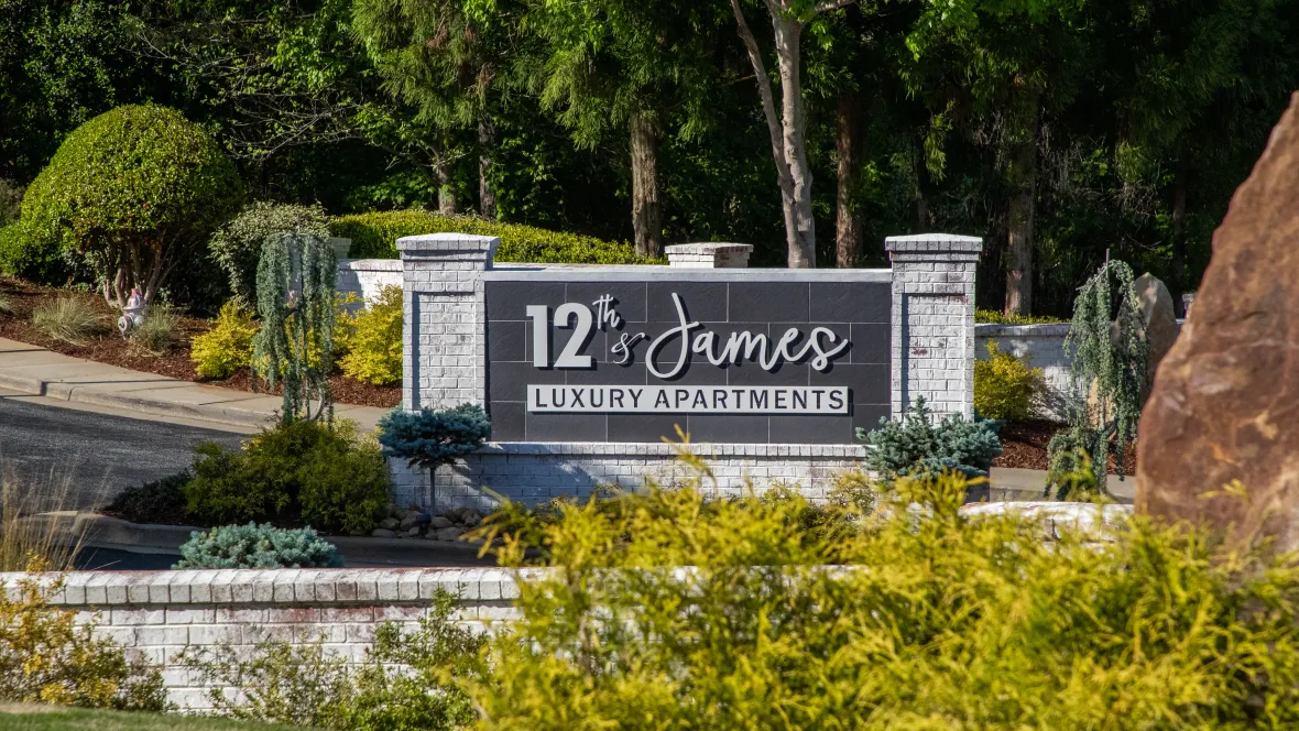 The front entrance sign that reads: "12th & James Luxury Apartments", surrounded by lush landscaping.