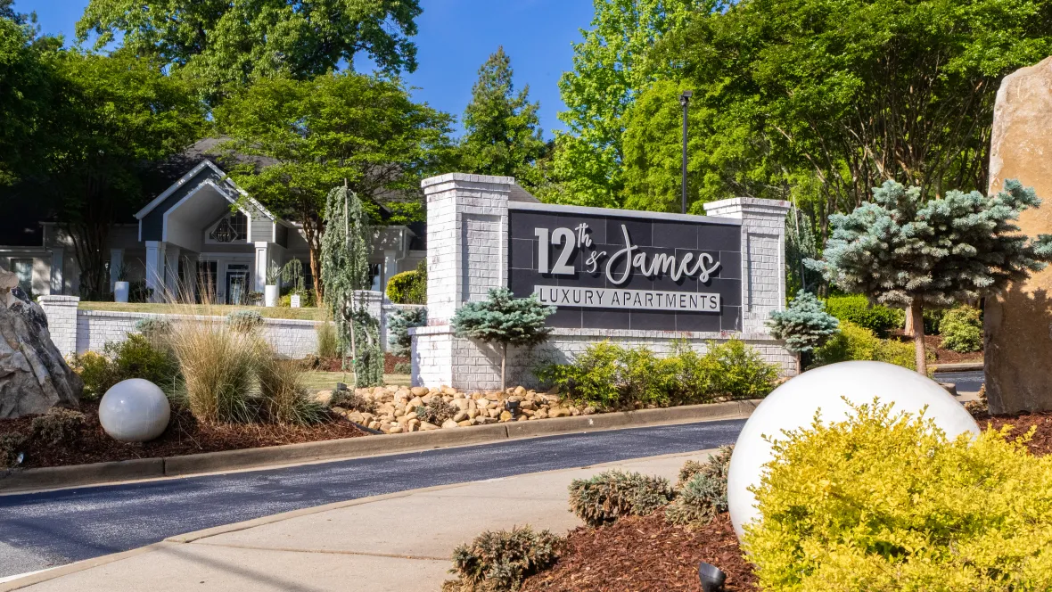 An angled photo of the front entrance sign that reads: "12th & James Luxury Apartments", surrounded by greenery and a gated entry in the background.