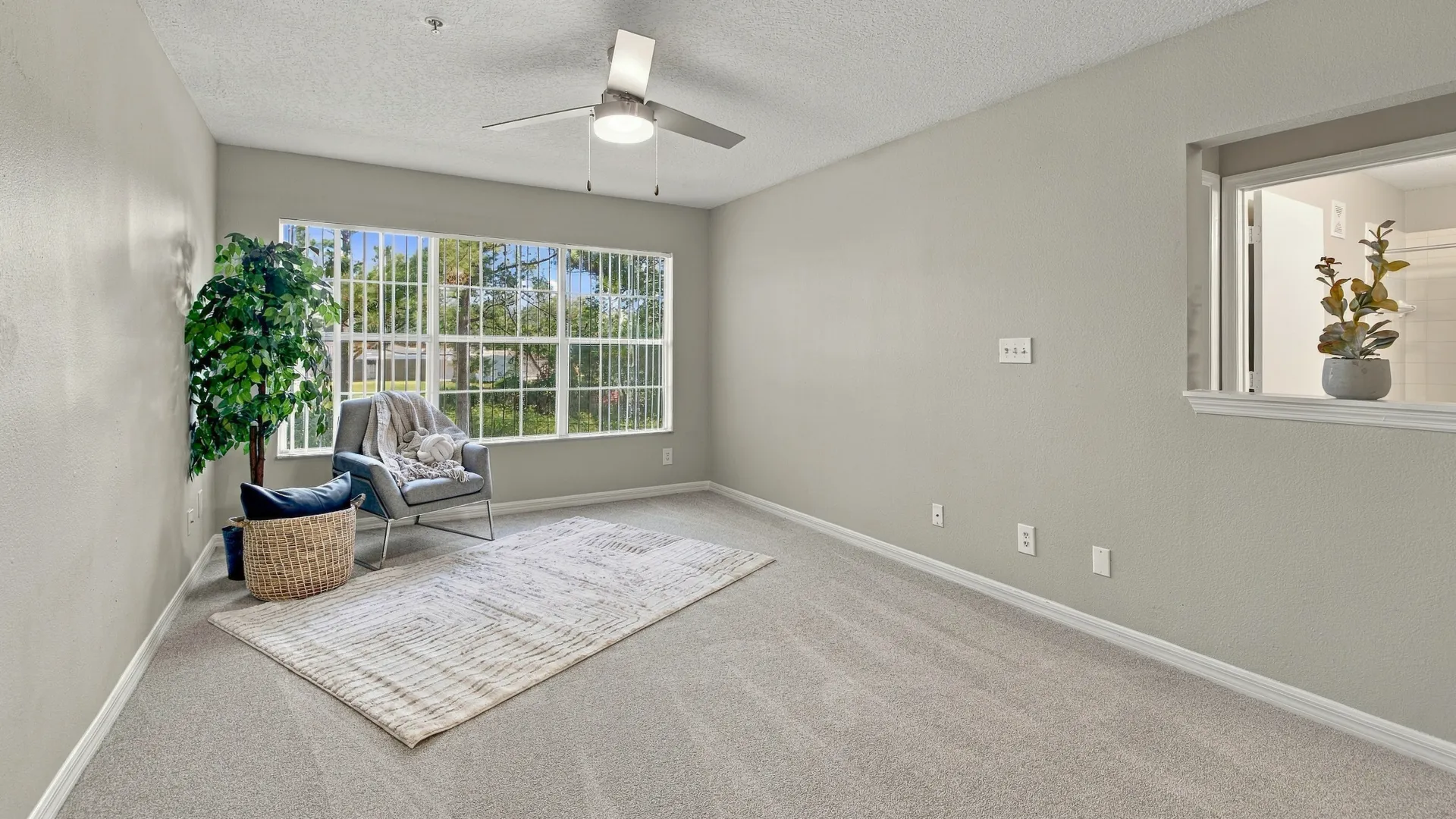 Living room at Eagle's Pointe featuring plush carpeting, a large window with vertical blinds, a ceiling fan, and a spacious open layout filled with natural light.
