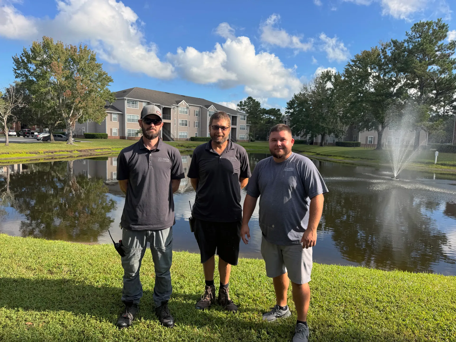 Three Eagle’s Pointe maintenance team members standing beside the community pond and fountain with apartment buildings in the background.