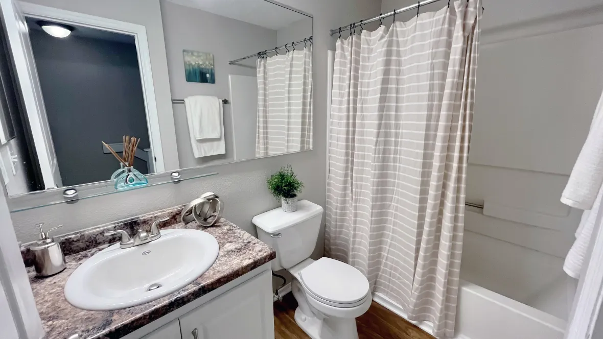 A brightly lit bathroom with granite-style countertops, an expansive mirror, elegant vanity lighting, and an oversized shower/tub combo.