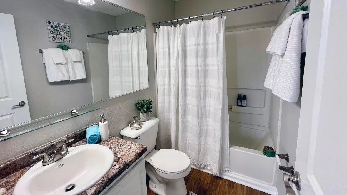 A bright bathroom featuring a shower/tub combo with a pristine white surround, large vanity, and an extensive, humongous mirror.