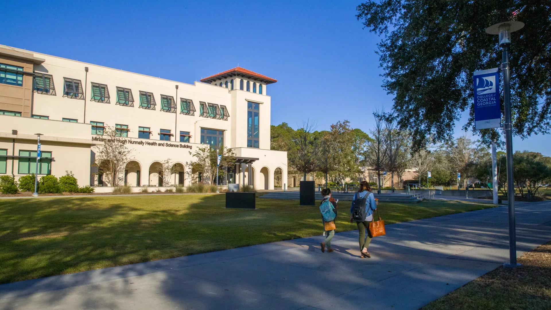 Exterior view of the College of Coastal Georgia campus with students walking along a sunny pathway and the Health and Science Building in the background.