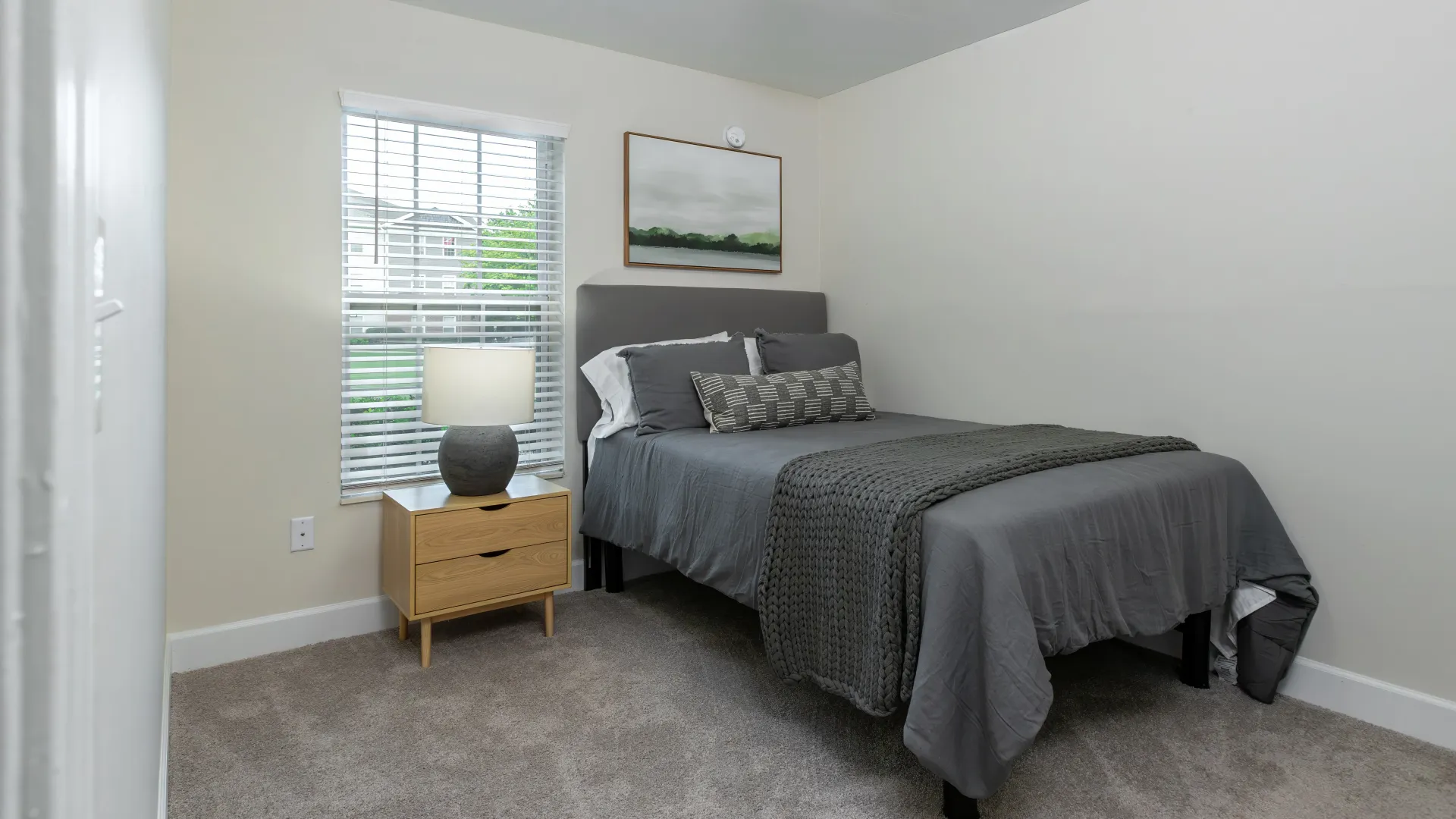 Modern bedroom with a gray bedspread, decorative pillows, and a wooden nightstand with a lamp.