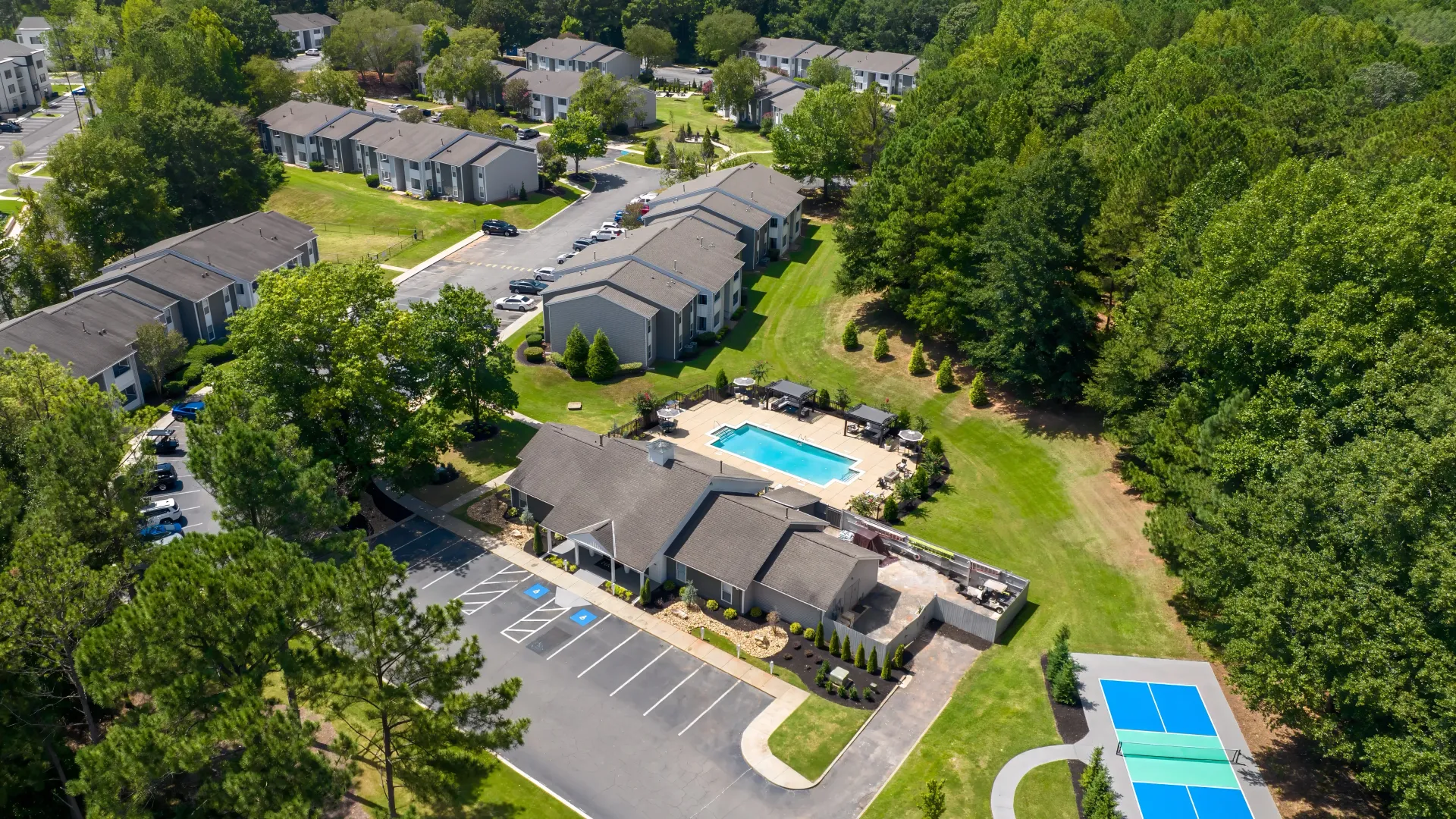 An expansive aerial view showcasing the robust community featuring the lush trees surrounding creating a serene ambiance, the exquisite expansive pool deck, and the vibrant pickleball court, offering an idyllic oasis to call home.