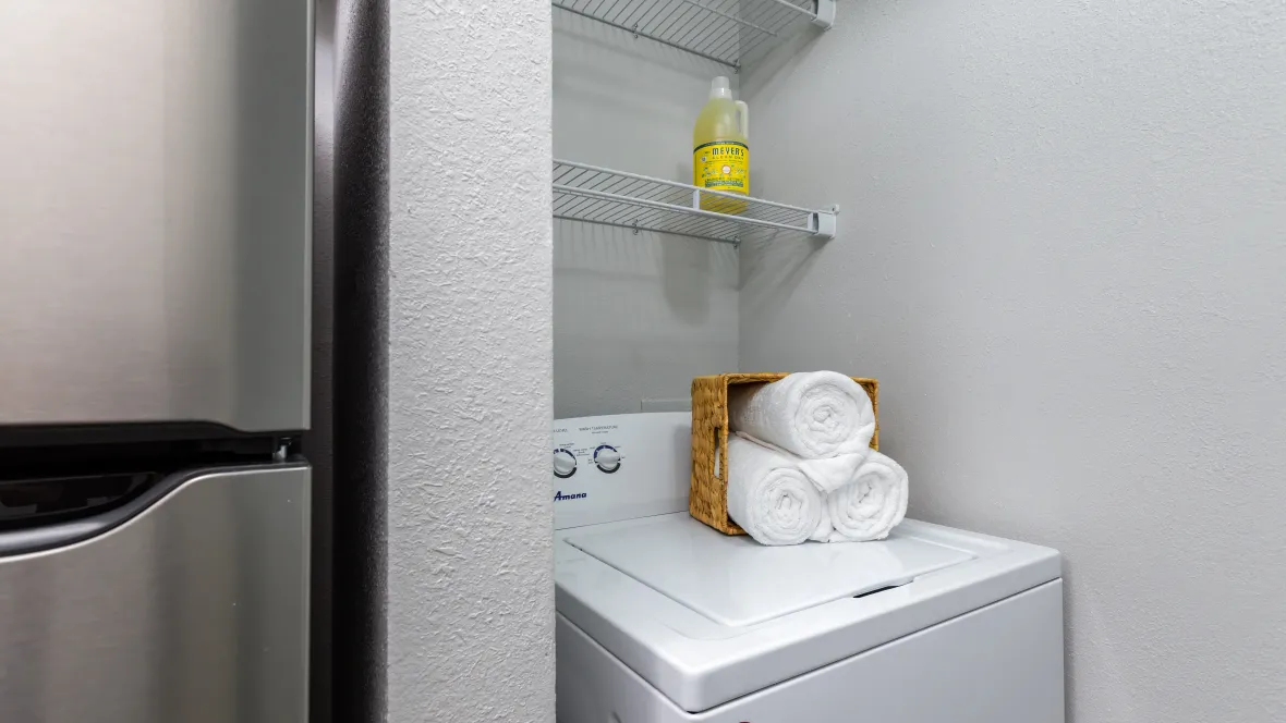 A top-loading washing machine situated conveniently near the kitchen with two rows of open-wire custom shelves above, providing versatile storage in the laundry space.