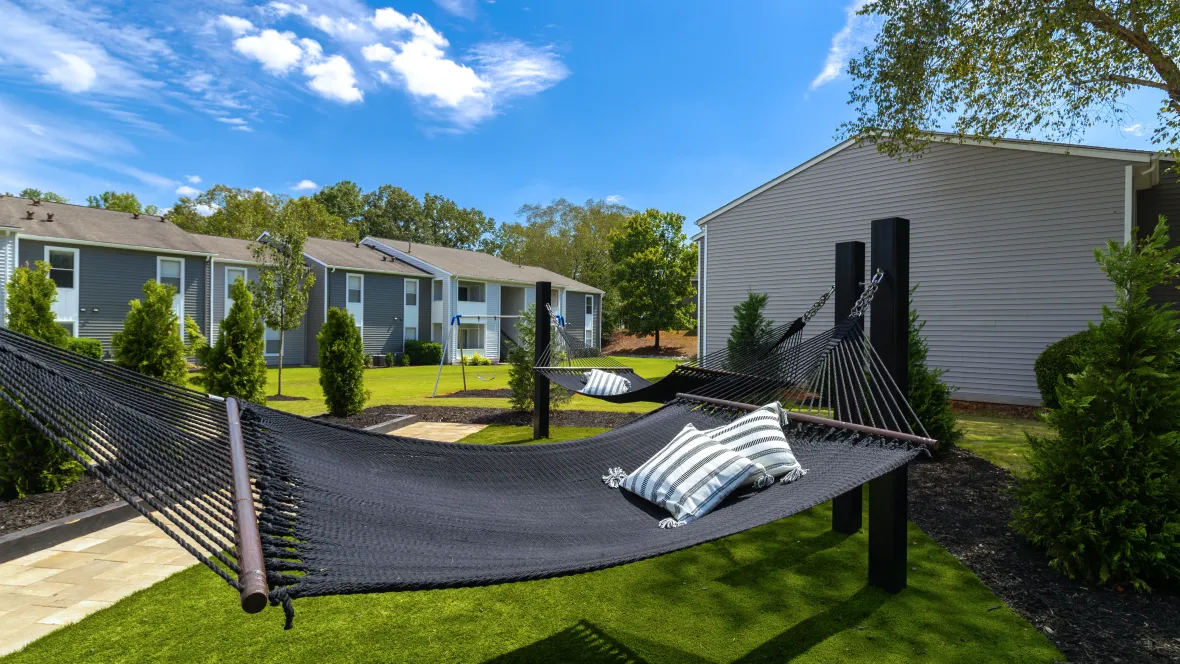 A paved walkway leading to a serene hammock garden with two loungers in a grassy oasis in a courtyard setting, offering a relaxing outdoor experience.