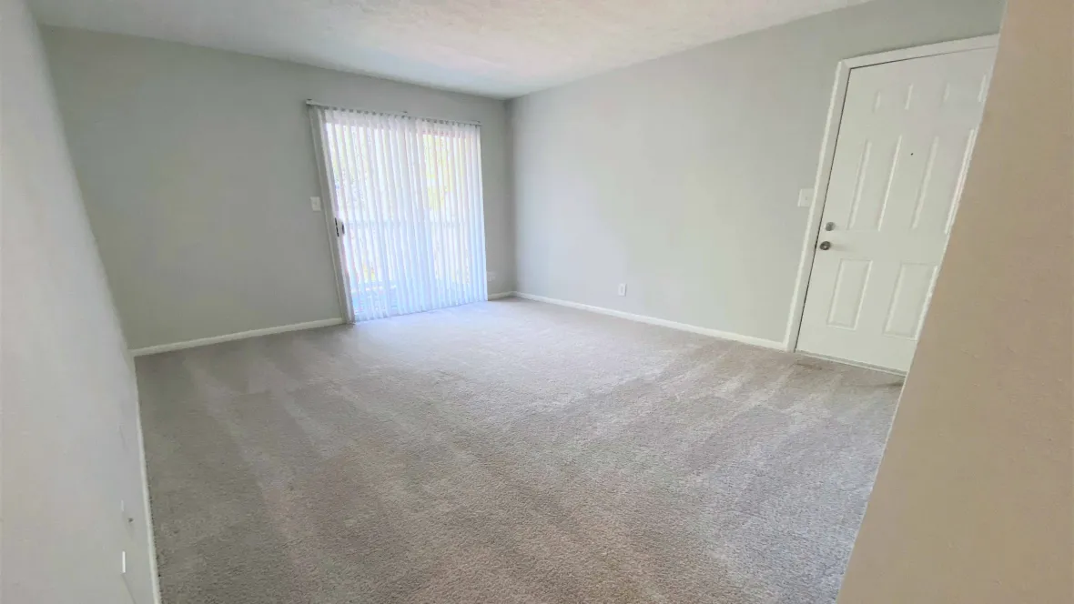 A cozy and spacious living room with plush carpeting and adjustable blinds on patio sliding doors.