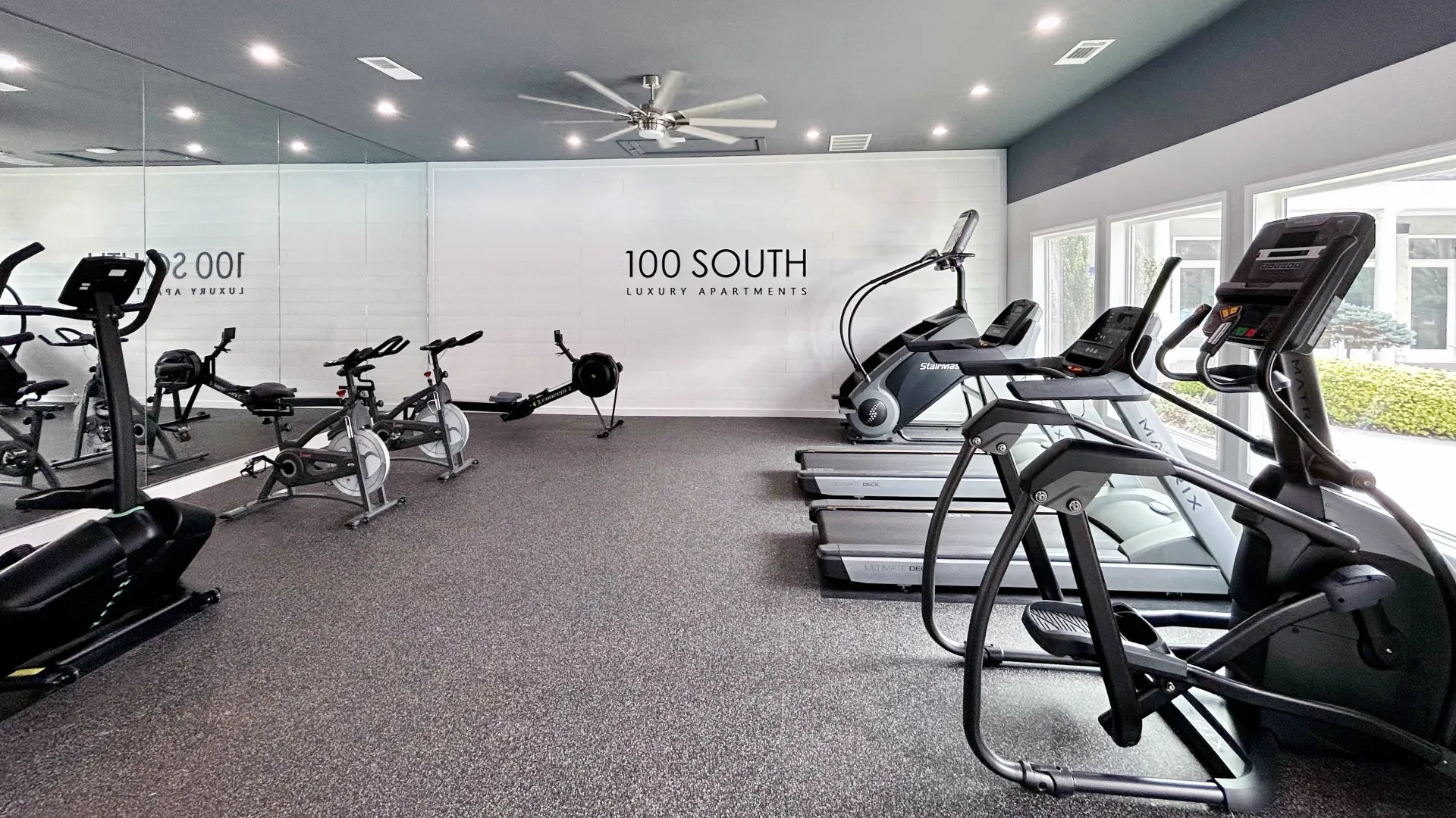 Modern fitness center at 100 South Luxury Apartments with treadmills, ellipticals, spin bikes, and rowers, surrounded by mirrors and windows offering ample natural light.