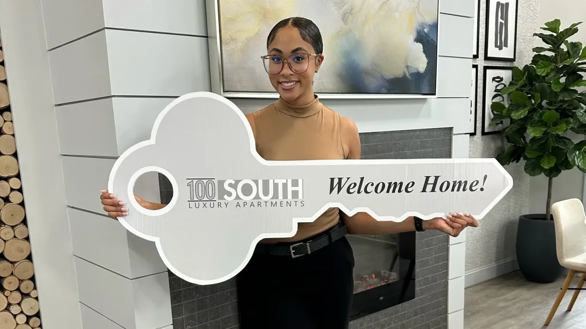 A woman in a tan shirt holding a sign in the shape of a large key that reads, "100 South: Welcome Home!"
