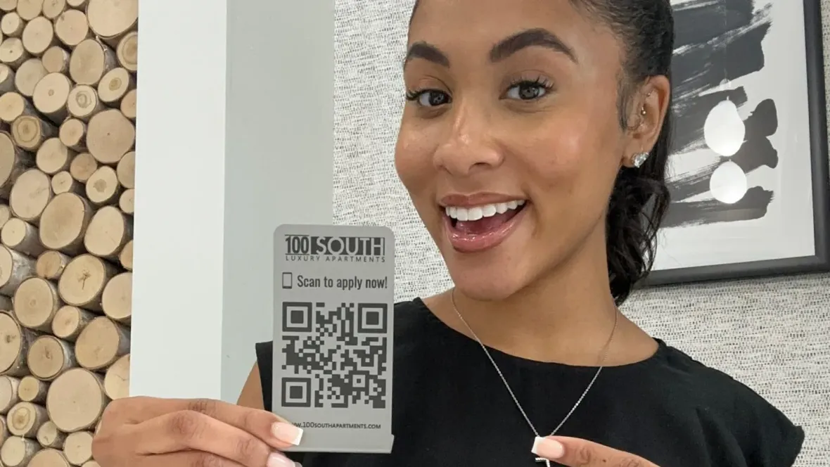 A woman in a black shirt holding a small metal sign with a QR code that says, "Scan Here to Apply Now!"