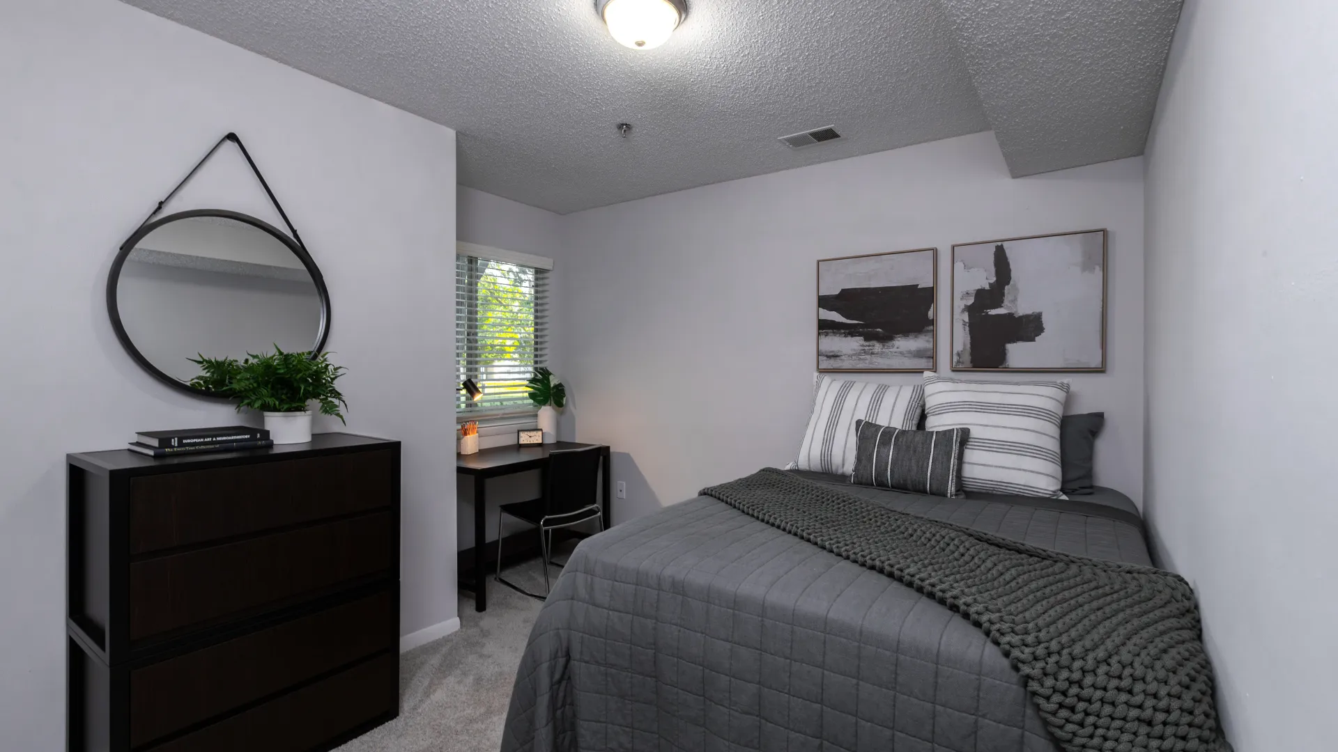 Contemporary bedroom with a gray bed, dark wood furniture, a round mirror, a plant, and a desk by the window.