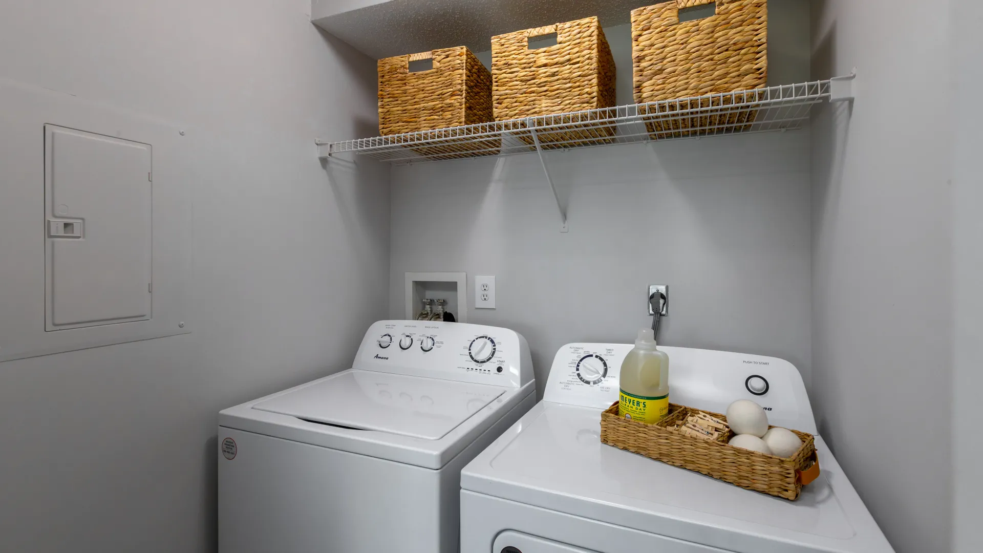 In-unit laundry room with a modern washer and dryer, wire shelving with wicker baskets for storage, and laundry essentials neatly organized.