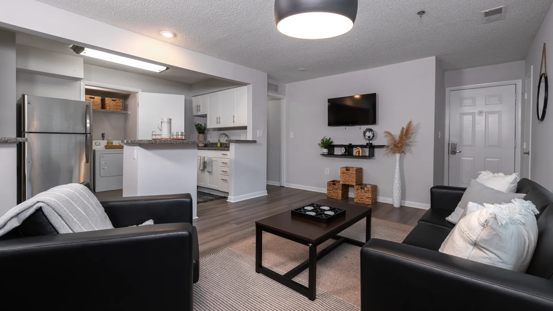 Open-concept living space with black leather seating, wall-mounted TV, modern kitchen with stainless steel appliances, granite countertops, and in-unit laundry area.