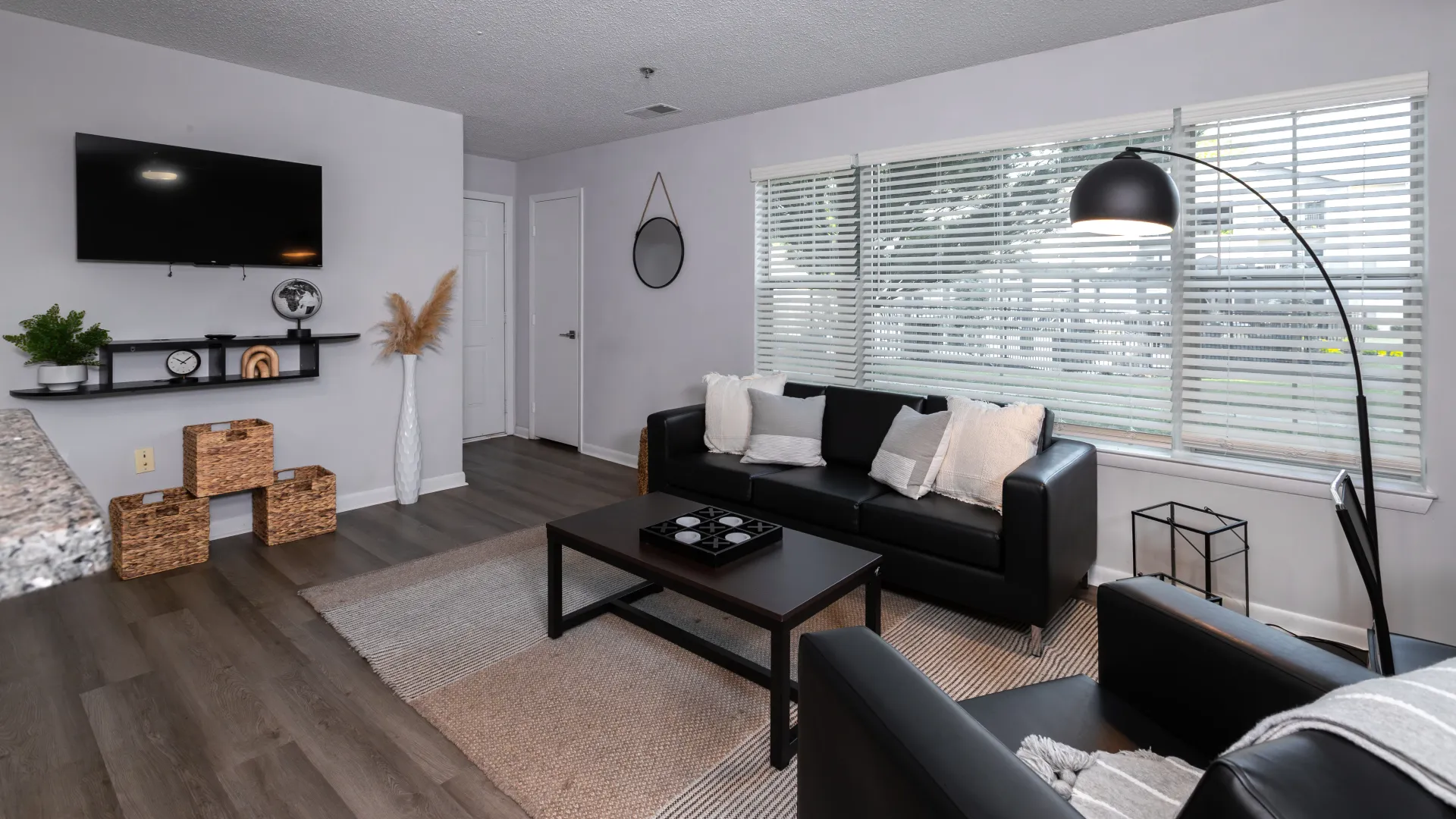 Bright living room with black leather seating, wall-mounted TV, coffee table, large windows with blinds, and stylish decor including wicker baskets and a floor lamp.