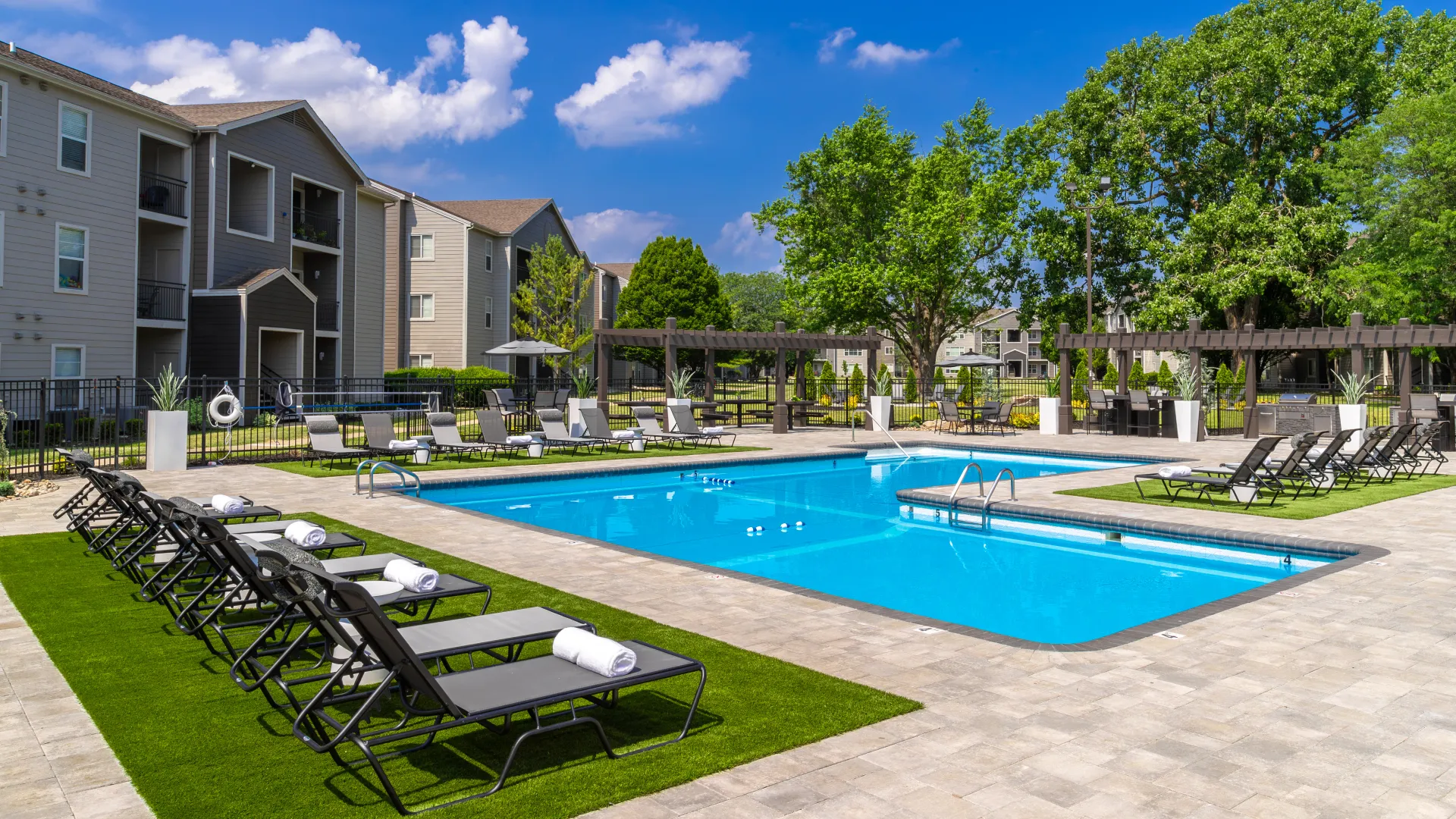 Poolside area with a blue pool, surrounded by lounge chairs, shaded pergolas, grilling stations, and beautifully landscaped apartment buildings in the background.