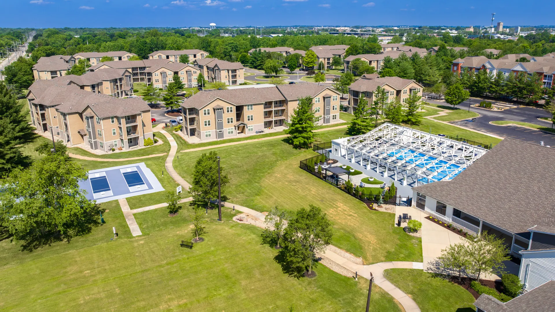Aerial view of The LINC Apartments community in Urbana, IL, showcasing the apartment buildings, pickleball courts, swimming pool area, and lush green surroundings.
