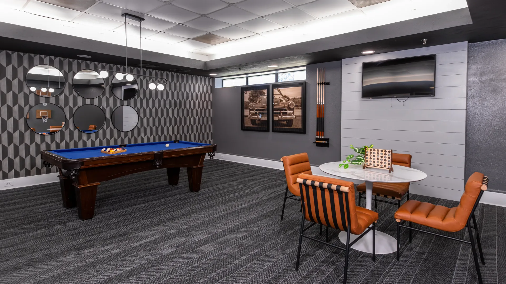 Game room featuring a blue felt billiards table, a round table with orange chairs, and modern decor with circular mirrors and vintage car photos on the walls.