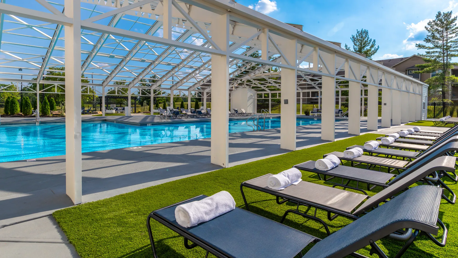 Pool deck featuring a row of loungers with rolled towels beside a clear blue pool under a modern, partially enclosed structure.