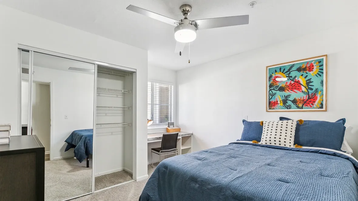An inviting apartment bedroom showing as fully furnished with a stylish-contemporary lighted ceiling fan with plush neutral carpeting with closet doors open to showcase customized shelves. 
