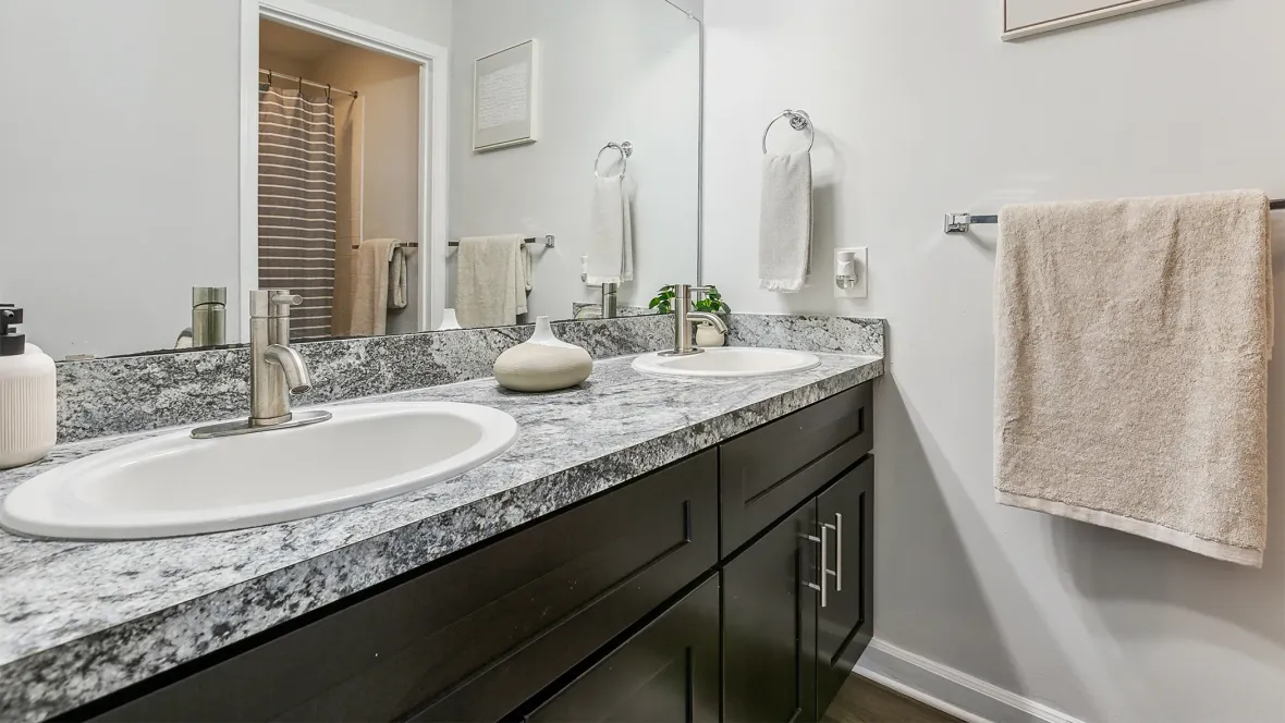 A meticulously designed interior bathroom featuring spacious granite-inspired countertops with dual sinks, practical under-cabinet storage, offering a view of walk-in showers reflected in large mirrors, complemented by strategically placed towel holders.