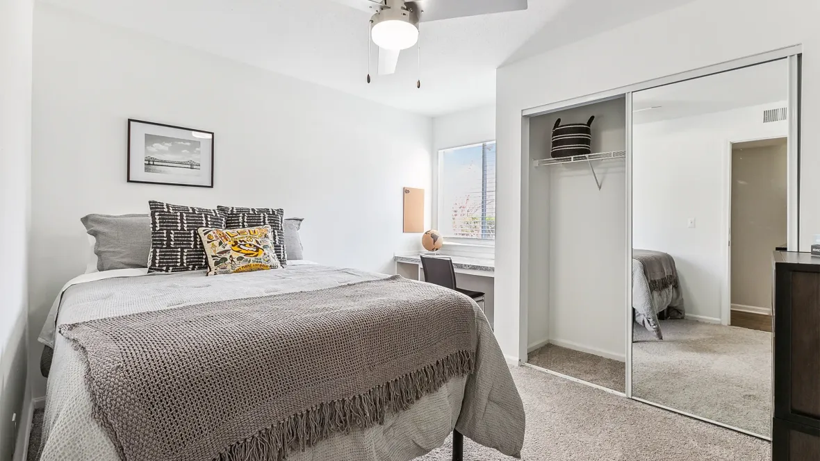 A spacious apartment bedroom with a full-size bed, a well-appointed desk space by the window, and mirrored sliding closet doors revealing a spacious closet with shelving.