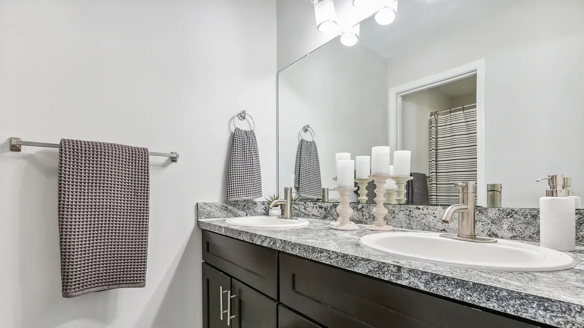 An exquisite bathroom, a realm of grandeur with rich dark brown cabinets providing ample storage beneath the generously sized grey granite countertop, all enhanced by the brilliance of a well-lit vanity.