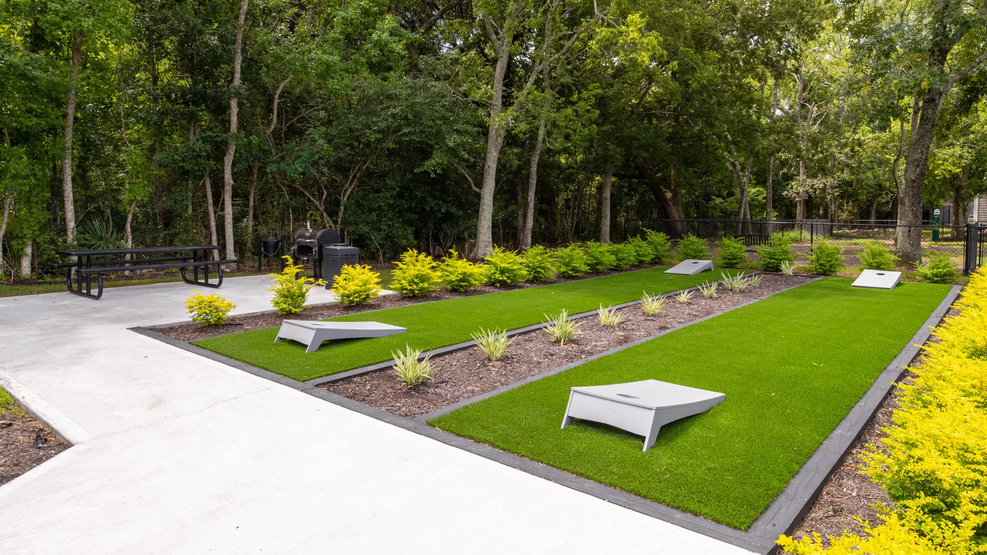 Cornhole boards set on green artificial turf, with picnic tables and BBQ grills in the background, surrounded by lush trees.