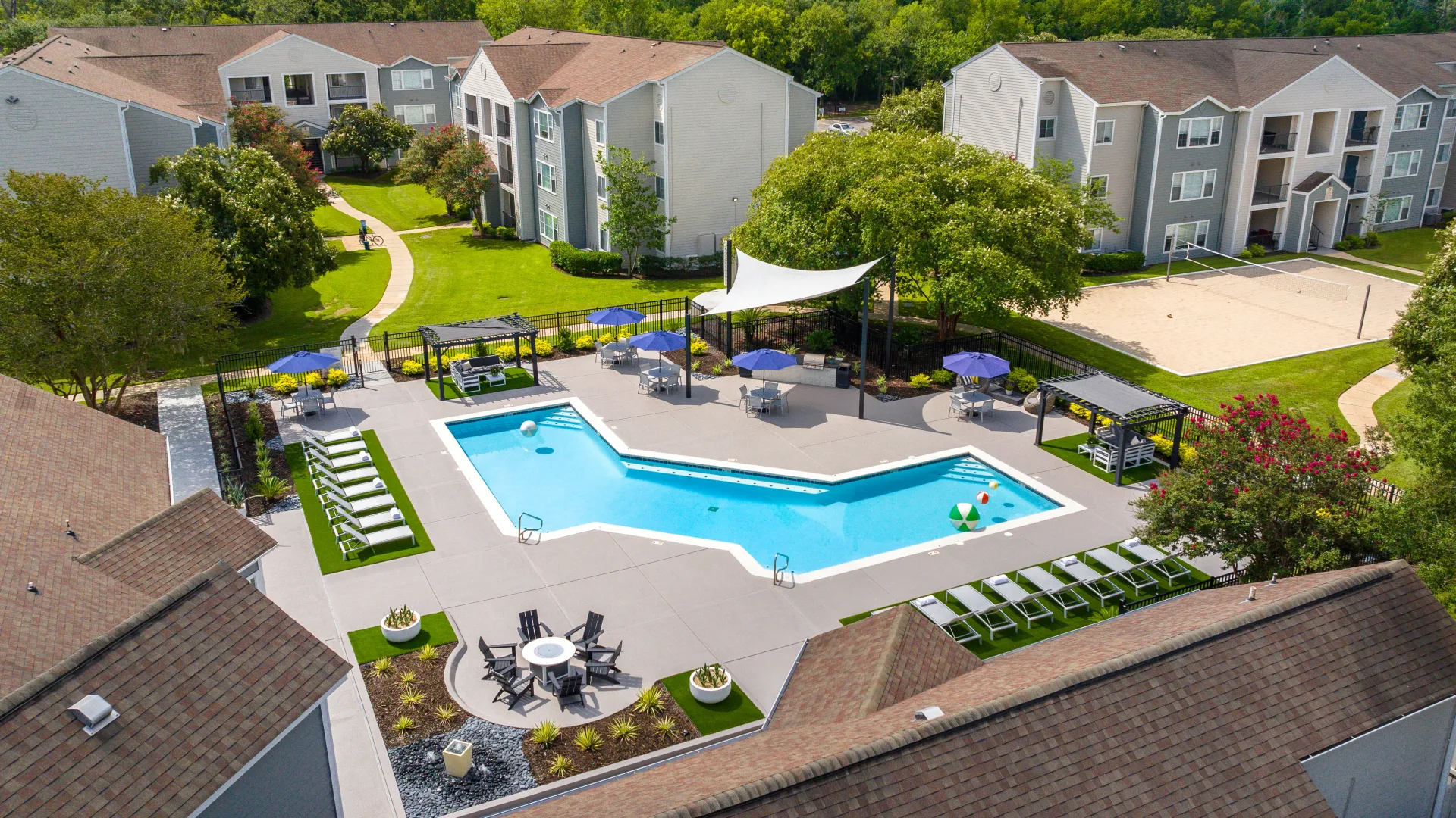 Aerial view of the pool area at Maison Burbank, featuring lounge chairs, shaded seating, a fire pit, and a volleyball court surrounded by green lawns and apartment buildings.