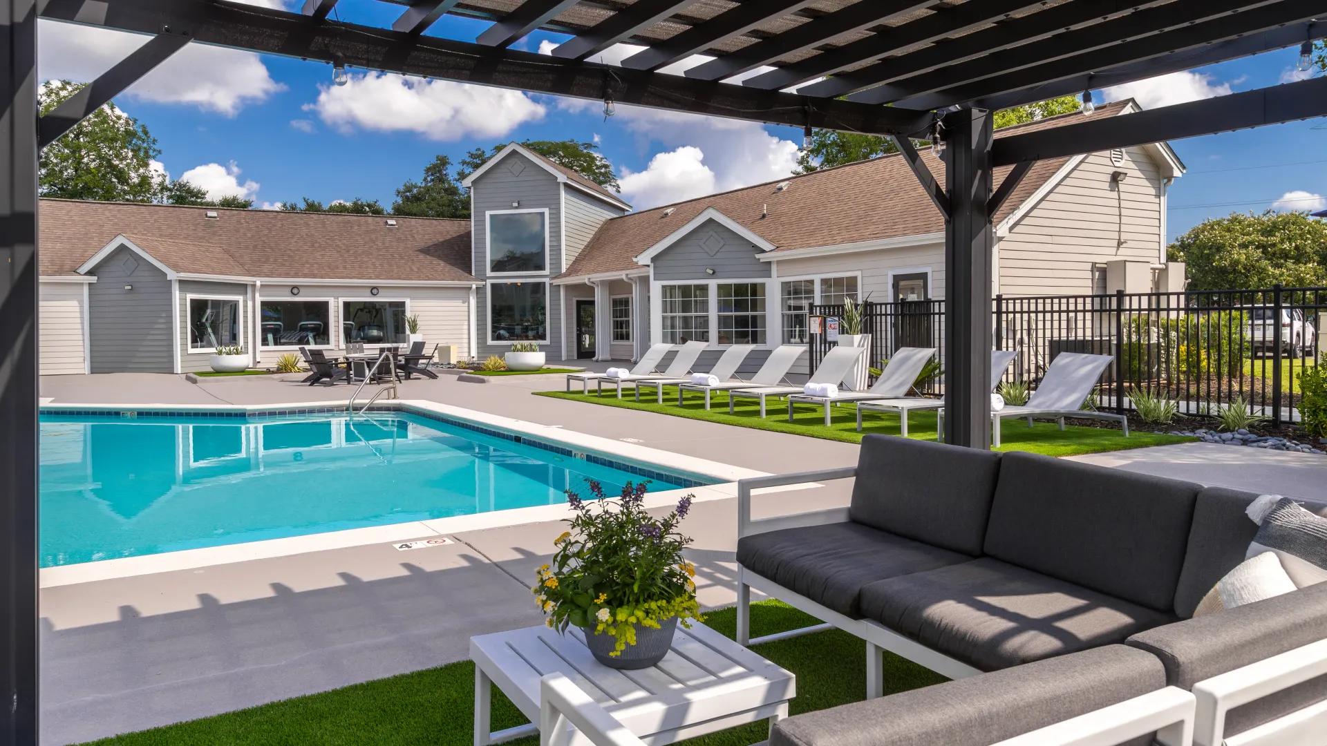 Pool area with lounge chairs, shaded seating under a pergola, and the clubhouse in the background under a blue sky with white clouds.