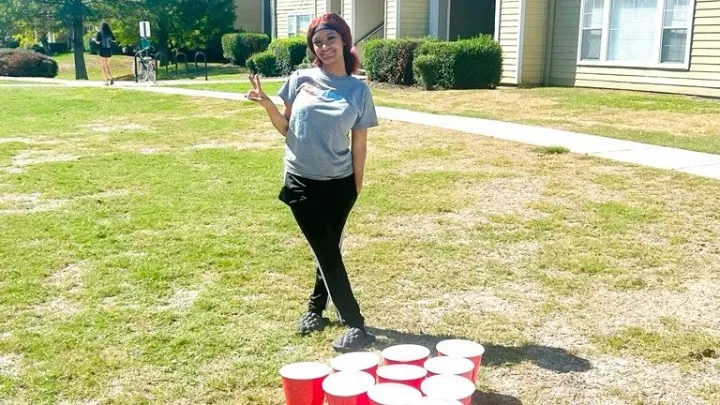 A resident standing in the courtyard in front of a jumbo yard pong game making a peace sign with their fingers.