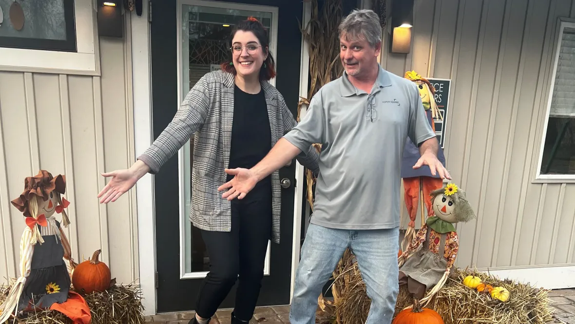 Two employees standing outside the leasing office with their arms out surrounded by beautiful fall decor.