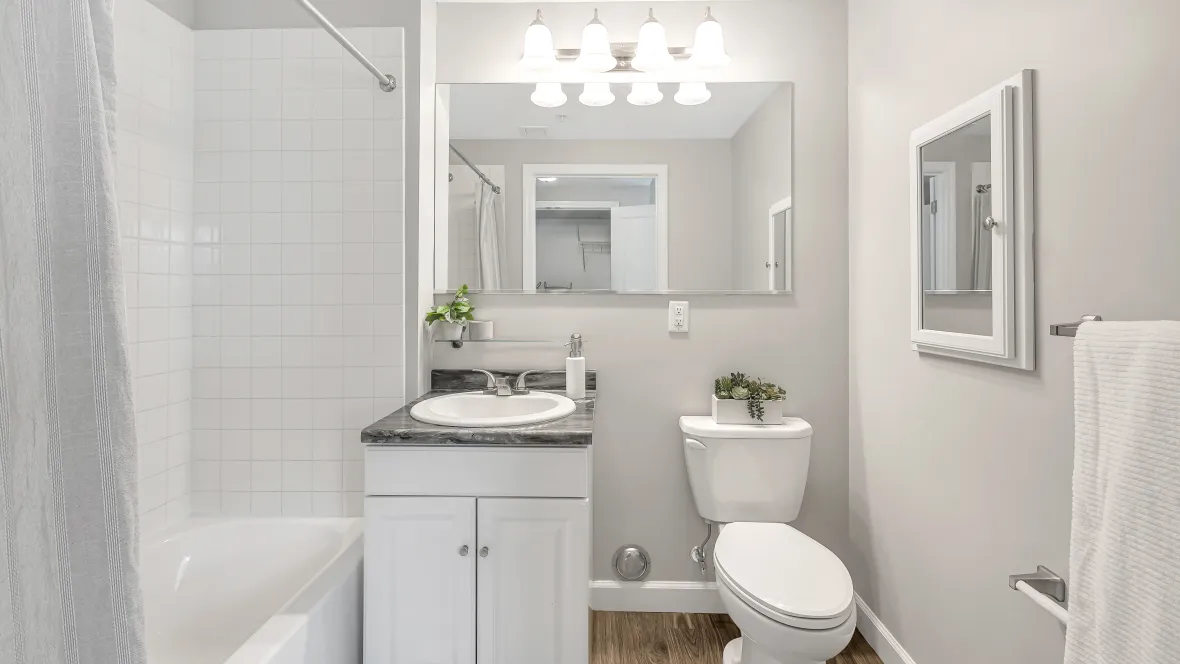 A contemporary bathroom with white tile-surrounding the sides of a shower/tub combo, ample lighting and large mirror over the sink, and a mirrored medicine cabinet on the wall to the right of the toilet. 