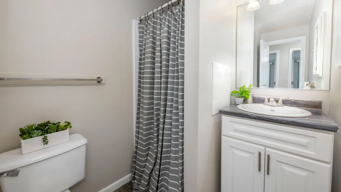 A bathroom with a walk-in shower left of the sink and a convenient towel bar near the shower. The countertop is black-fusion granite style, and the cabinetry is white.