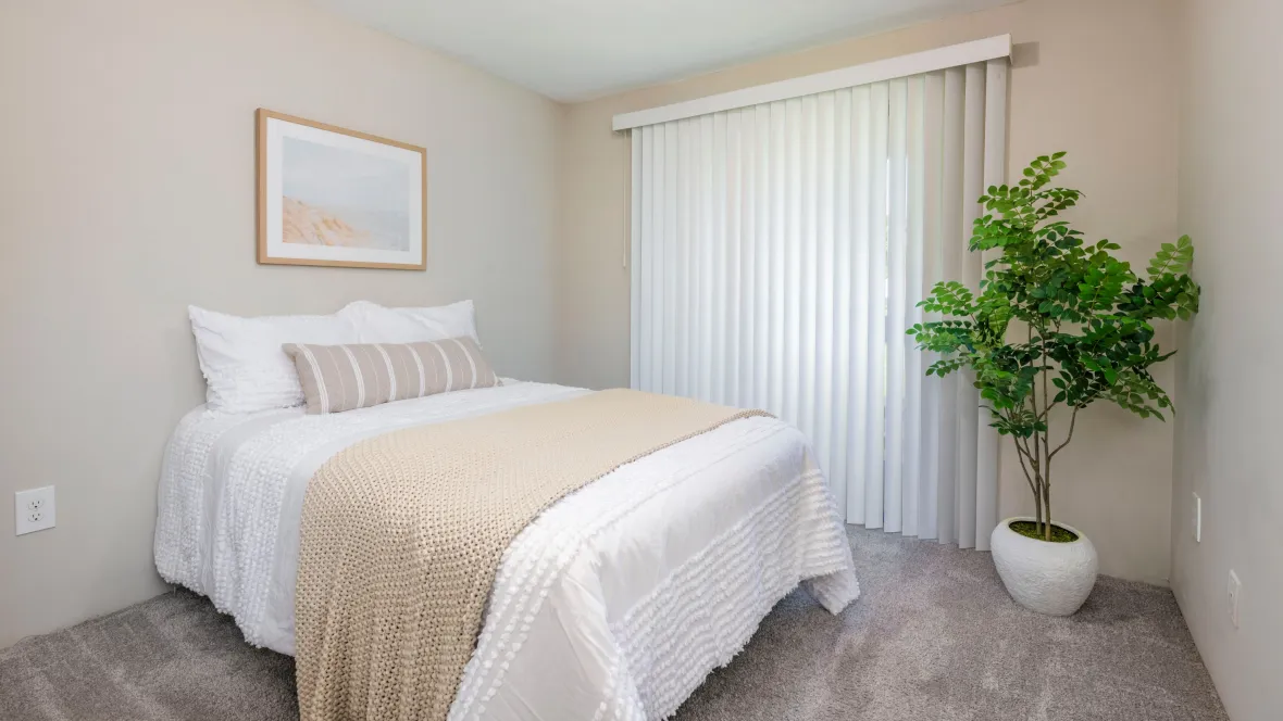 A bedroom with plush carpeting and large slider with wood-style blinds leading to a private patio.