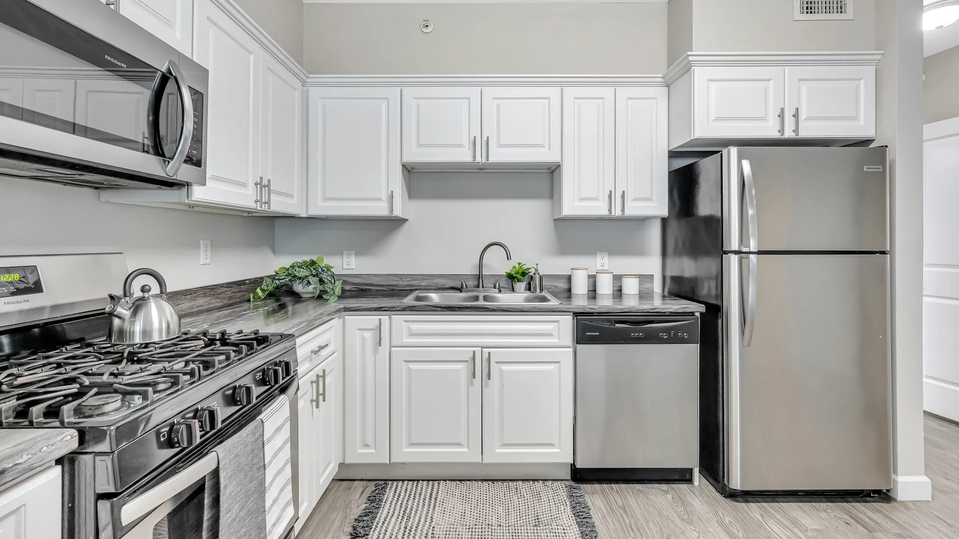 A modern, well-lit kitchen with stainless steel appliances, white cabinets, black-fusion granite-like countertops, wood-like flooring, ample storage, fridge, stove, dishwasher, microwave, and a double sink.