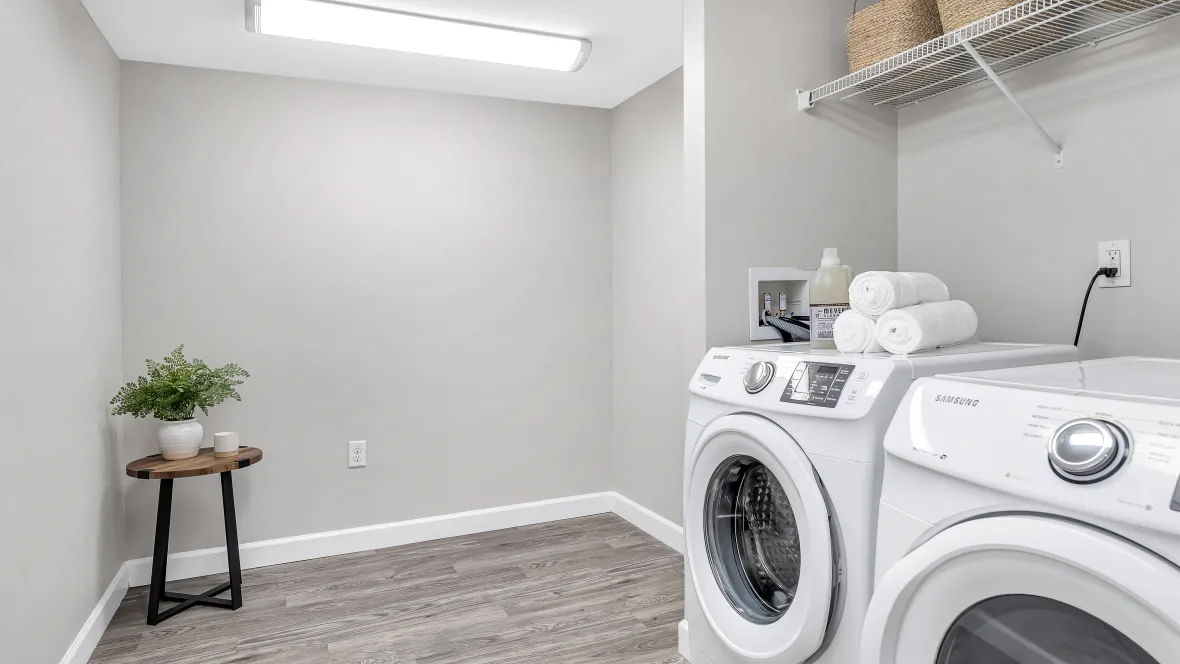 A well-lit and very spacious laundry room with full-size washer and dryer appliances and open wire shelving overhead for storage.