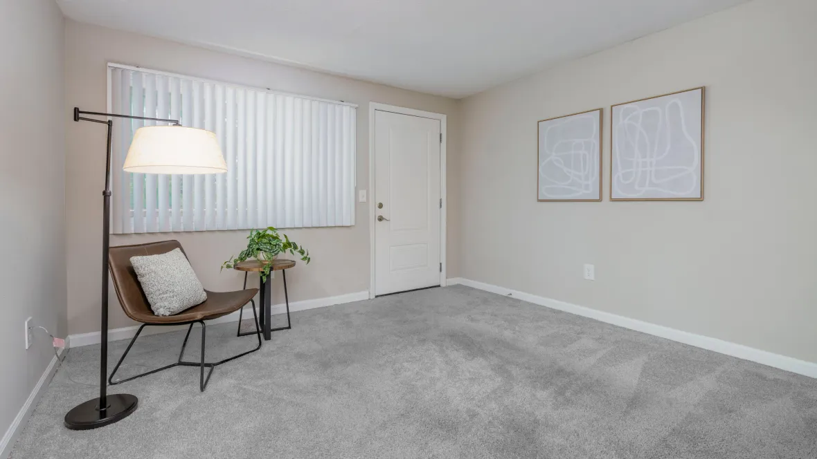 Large living room space with plush carpeting and a large picture window with adjustable blinds.