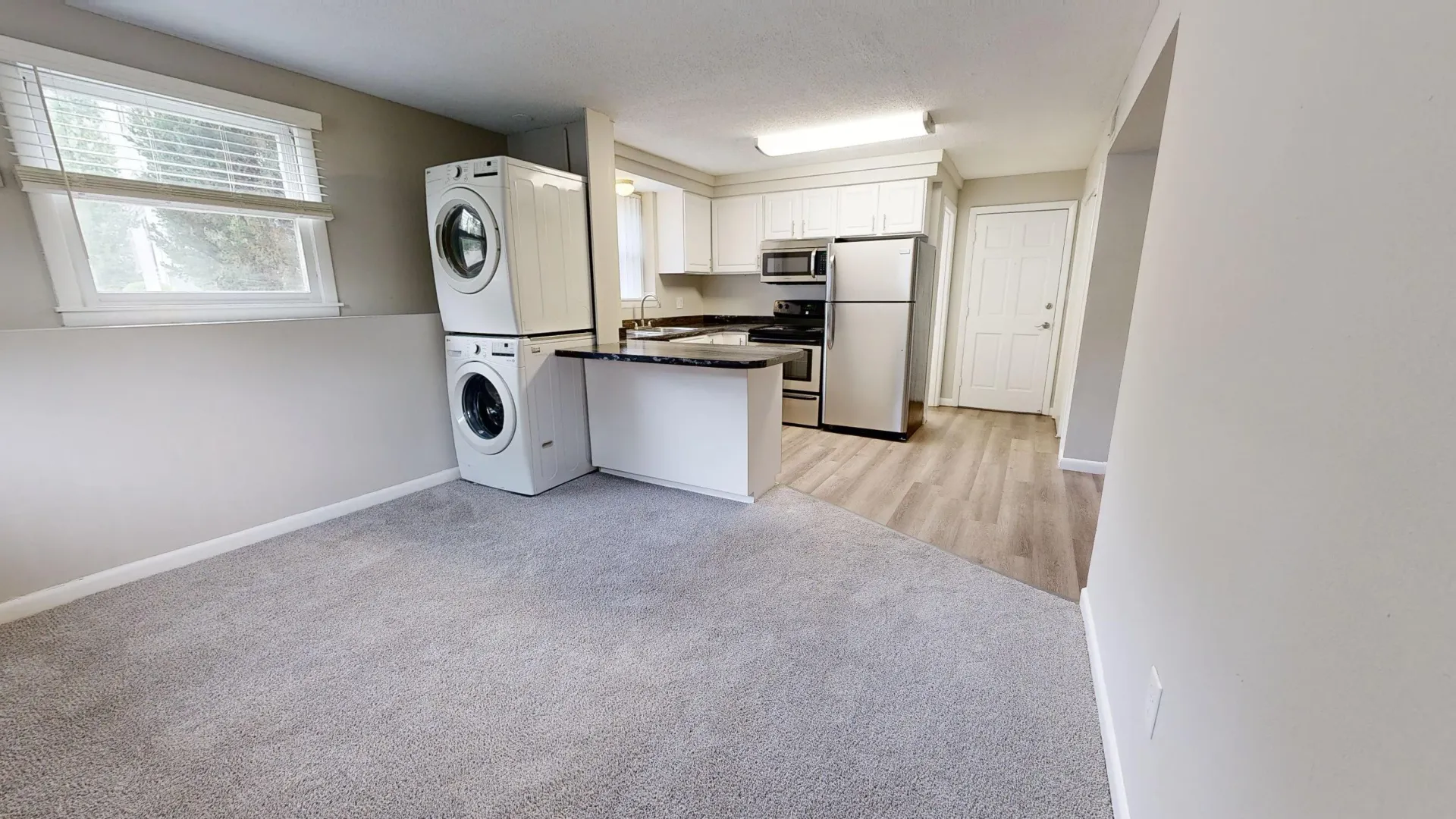 Open-concept living space in The Valley floor plan at Alpine Commons Apartments with modern kitchen, stainless steel appliances, in-unit washer and dryer, and carpeted living area.