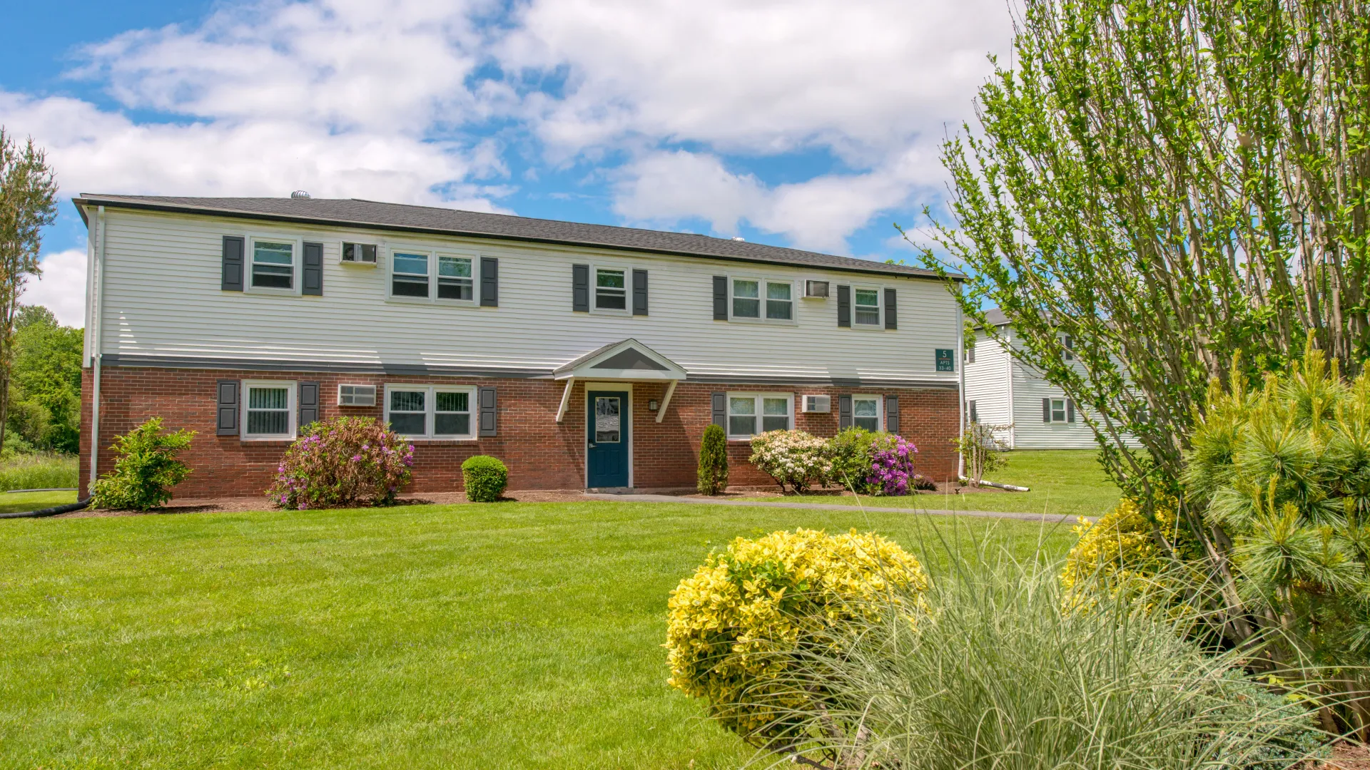 Aspen Chase apartment building surrounded by landscaped lawns, flowering bushes, mature trees, and a bright blue sky.