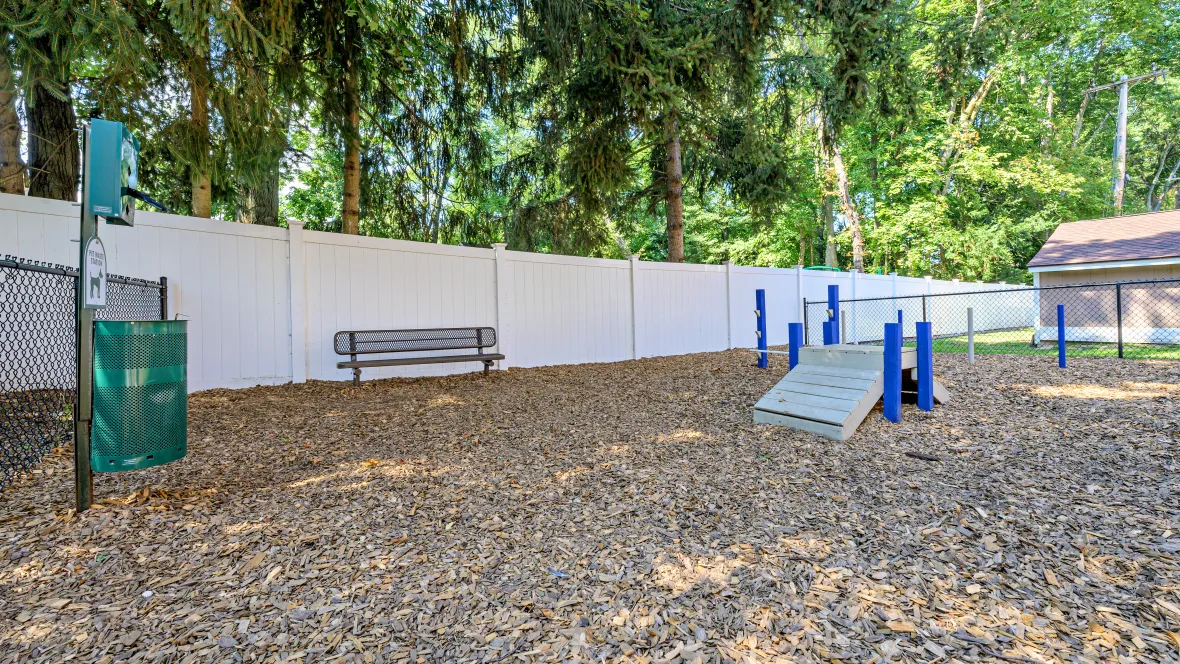 Our off-leash dog park covered in wood-chip mulch featuring agility equipment, a bench, and a waste station.