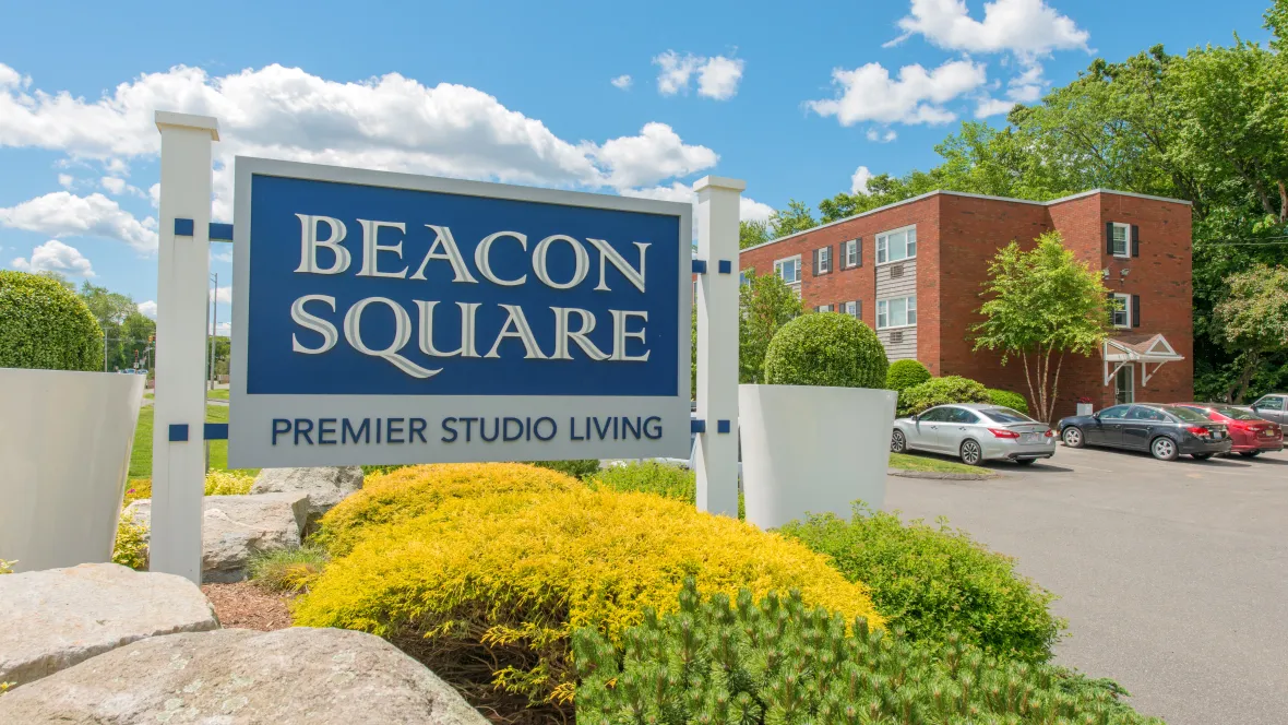 A photo of the vibrant community sign that reads "Beacon Square Premier Studio Living" surrounded by lush greenery and boulders.