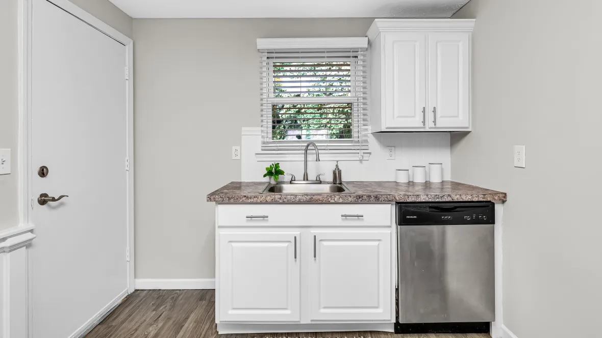 A corner of the kitchen that features a window over a small section of the granite-style countertop and white cabinetry with a stainless steel dishwasher built-in.