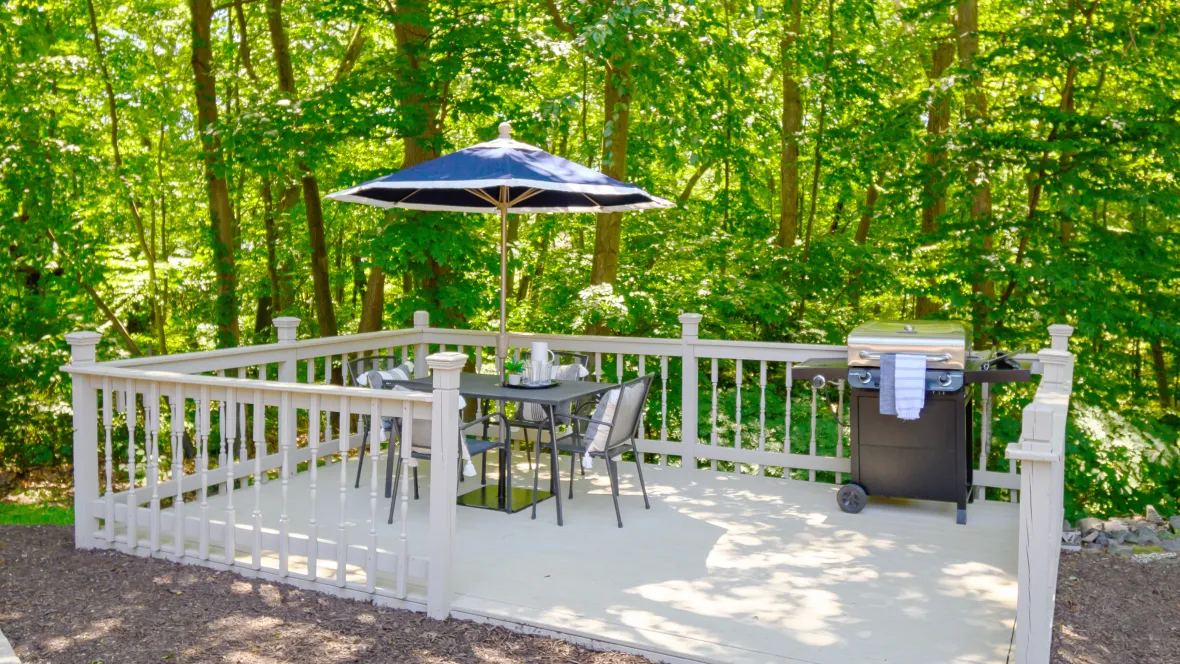 A white deck with a grill and picnic table with umbrella in a wooded corner of the apartment community.