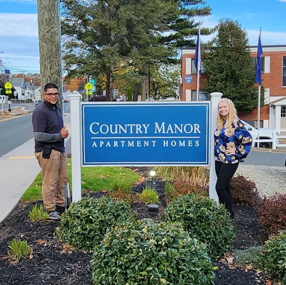 Country Manor Apartments team members standing beside the community entrance sign surrounded by professionally maintained landscaping and welcoming curb appeal.