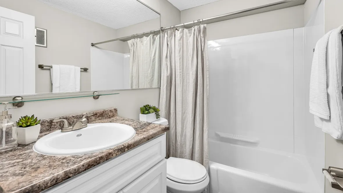 An elegantly designed bathroom with an extended mirror, white cabinetry, soothing vanity lighting, a shower/tub combo, and granite-style countertops, offering relaxation.